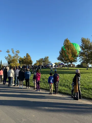 Families line a path in the morning sun as the balloon peeks through the trees ahead.