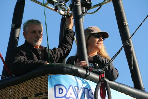 A man reaches for the burner controls while a woman in a cap watches from the basket.