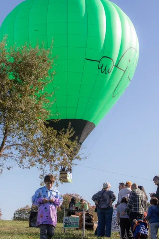 A small hot air balloon rises over a distant tree line as a few people watch below.