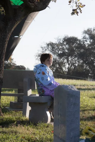 A person in a patterned costume shirt sits quietly on a bench in dappled shade.