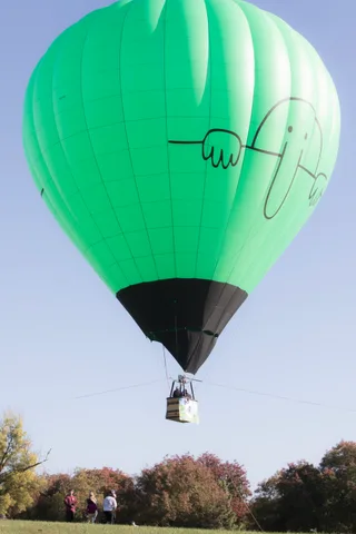A dark hot air balloon floats high above the tree line with tiny figures watching below.
