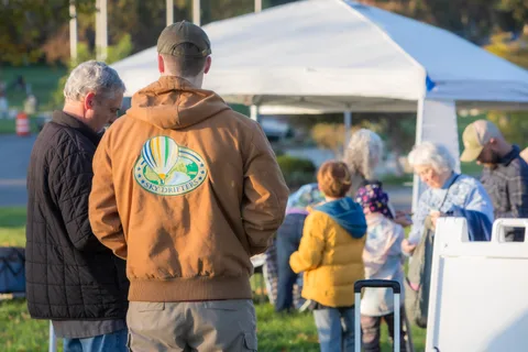 A man in a tan hoodie stands among a crowd near the event tents.