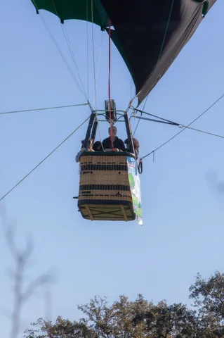 A rider waves from a hot air balloon basket rising directly overhead against blue sky.