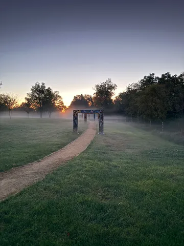 Golden morning light and mist glow over a park path leading to an arch at sunrise.