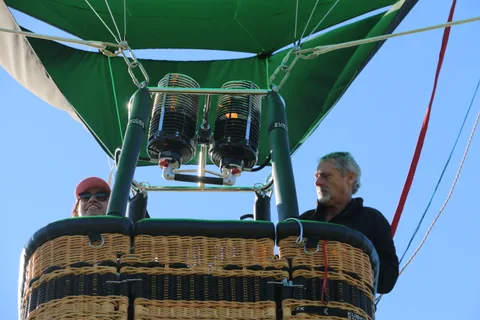 Two men in the balloon basket work near the burners under a clear sky.