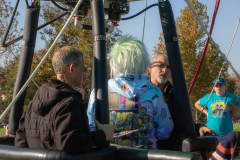 Two men, one in a green wig, stand near the balloon basket seen from behind.