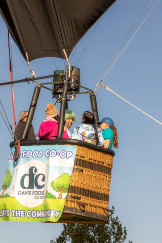 Riders lean over a hot air balloon basket bearing a full Davis Food Co-op banner, one waving down.