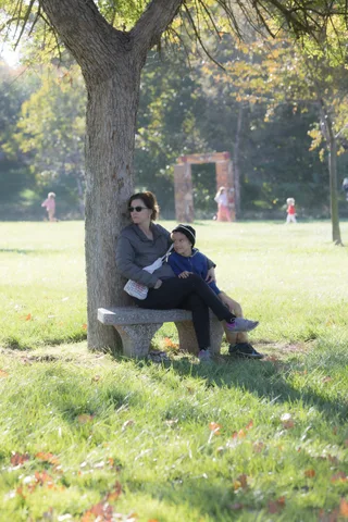 A woman and child sit together on a bench under a tree, a decorative arch in the distance.