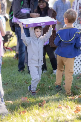 A child in a costume hoists a decorated box overhead while another child watches.