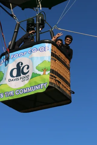 Two riders wave from the balloon basket above the sponsor banner.