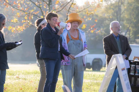 A woman speaks into a megaphone beside a costumed scarecrow character and a man in a blazer.