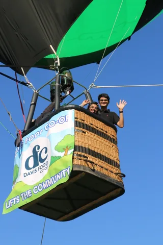 Riders wave from the balloon basket beneath a clean sponsor banner and blue sky.