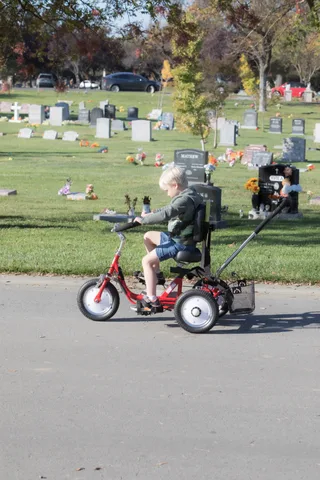 A child rides an adaptive tryke along a cemetery path as an adult walks alongside.