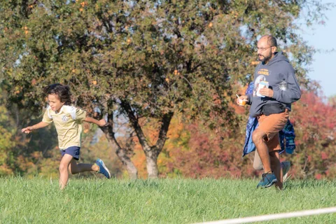 A boy runs through the grass with arms outstretched against the autumn trees.