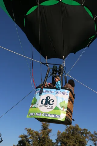 Riders peek over the balloon basket above the sponsor banner against blue sky.