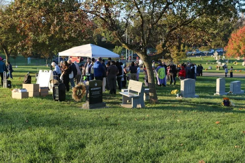 A small crowd gathers near a tent under autumn trees in the cemetery grounds.