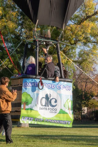 A man in a tan jacket claps near the hot air balloon basket.