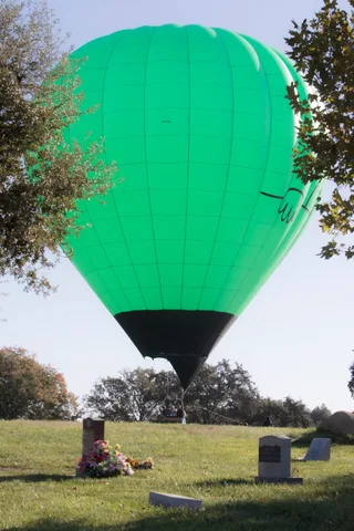 A hot air balloon descends low over the cemetery, gravestones with fresh flowers in the foreground.