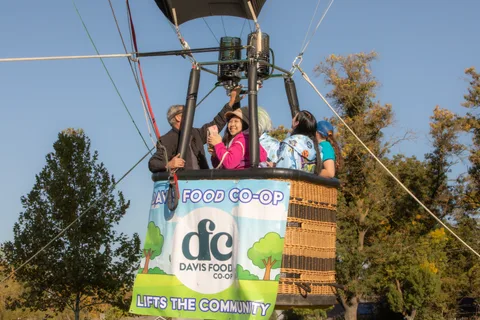 Riders stand in a hot air balloon basket above a Davis Food Co-op sponsor banner.