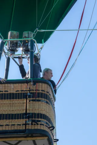 Two men operate the burner inside a hot air balloon basket against the sky.