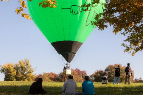 A hot air balloon rises over the trees as a small crowd watches from the field.