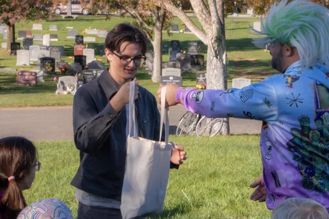 A man in glasses hands a tote bag to a costumed volunteer with teal hair.