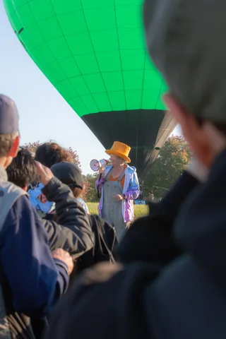 A presenter in an orange sun hat speaks into a megaphone as the balloon floats behind her.
