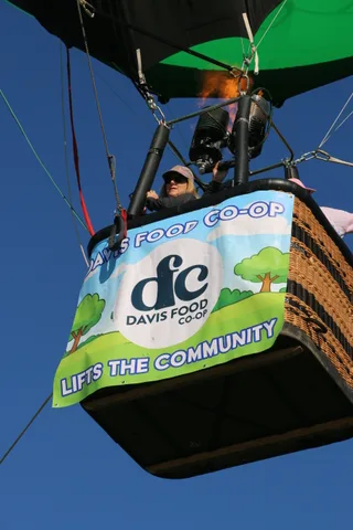 Riders wave from the balloon basket in bright sun above the sponsor banner.