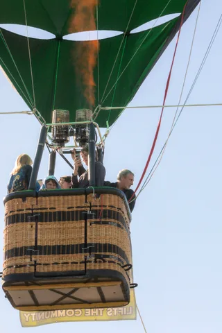 The underside of a hot air balloon basket shows the burner equipment and ropes, riders barely visible.
