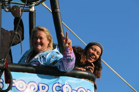 Two women laugh and wave from a hot air balloon basket in golden morning light.