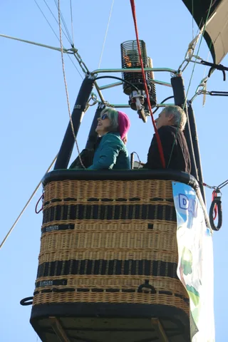 A woman with pink-streaked hair rides in the basket beneath the burner.