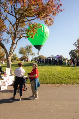 Two women stand on a paved path talking, a crowd and tent visible in the distance.