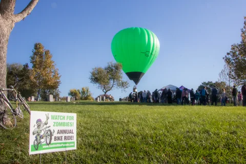 The hot air balloon floats alone above an open grass field lined with autumn trees.