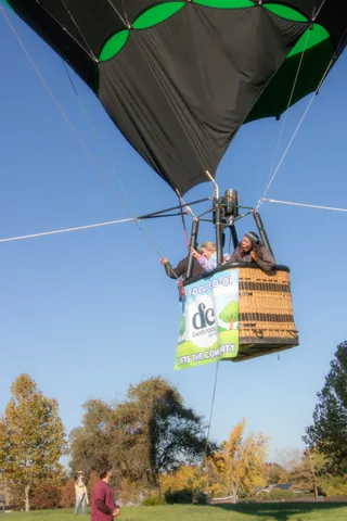 Riders wave from the balloon basket with rigging lines crossing the blue sky above.