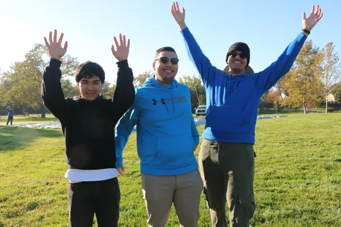 Three young men throw their arms up and cheer together on the sunlit lawn.