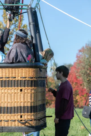 Two riders stand in a balloon basket on the ground with autumn trees behind them.