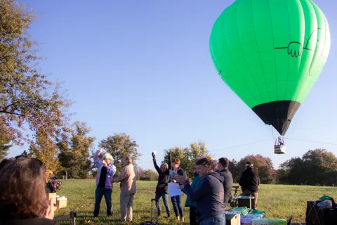 A green hot air balloon rises into the sky as a small crowd waves from among the headstones below.