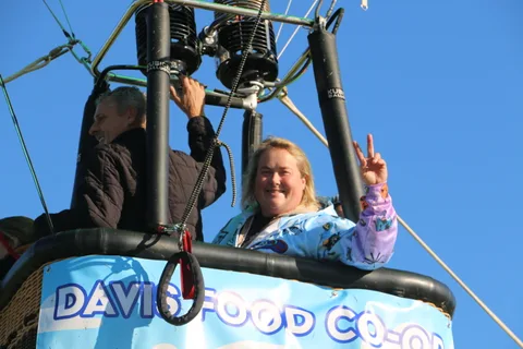 Two riders wave and flash a peace sign from a hot air balloon basket.