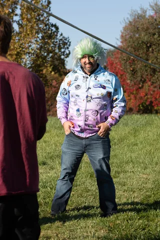 A man in a green wig and patterned zombie jacket stands candidly in the park.