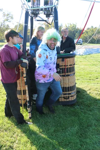 A group including a rider in a green wig gathers beside the balloon basket on the ground.