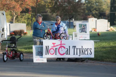 Two women stand with a trike near the NorCal Trykers trailer among headstones.