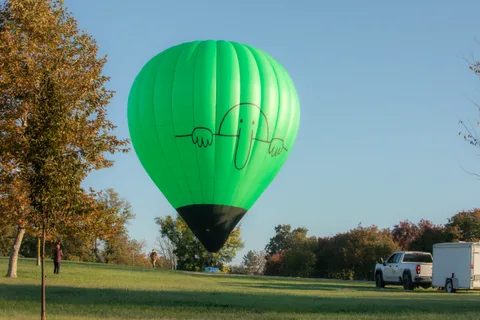 The balloon rests on the grass field with vehicles and trees in the background.