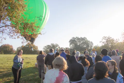 The balloon floats above trees as a seated crowd watches from the grass below.