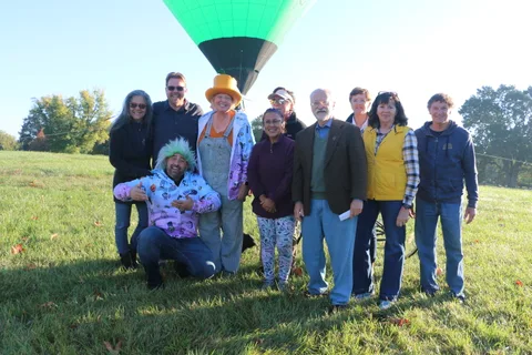 A group of organizers, one in a colorful wig, poses together on the lawn before the balloon launch.