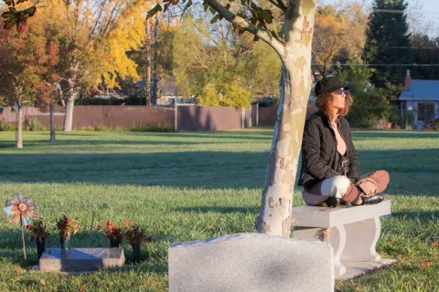 A woman sits cross-legged under a tree, sunglasses on, in quiet reflection.
