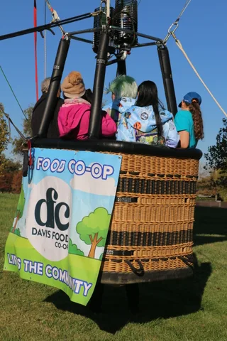 Costumed riders share the balloon basket above the sponsor banner.