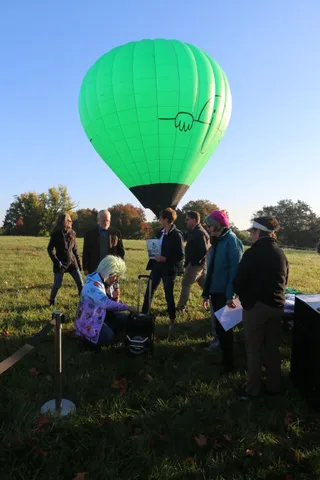 An emcee in a green wig speaks into a megaphone with the balloon rising behind him.
