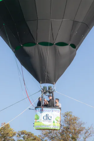 A hot air balloon basket carries riders into the sky above a sponsor banner reading Lifts the Community.