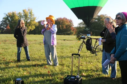 A woman in an orange top hat addresses a small crowd with the balloon towering behind her.
