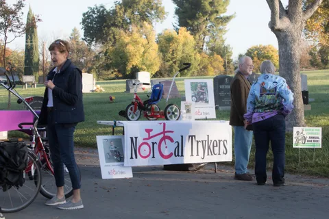 A woman stands near a trike and the NorCal Trykers sign in dappled shade.